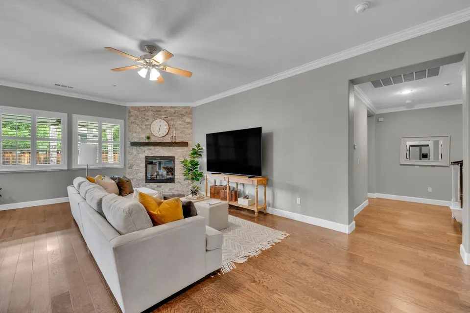 Family room with a stone-faced fireplace, ceiling fan and windows beside the open kitchen.