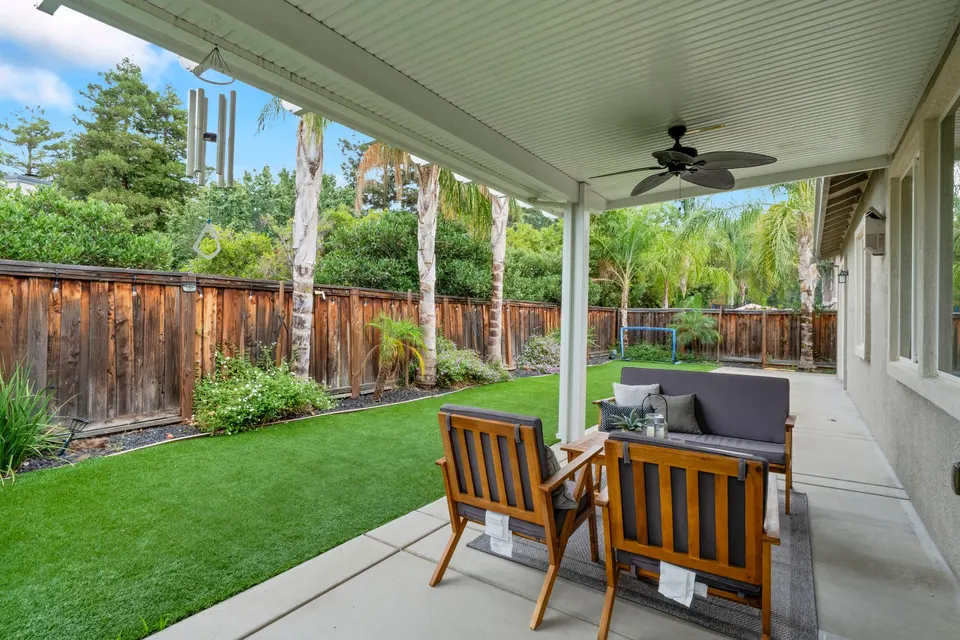 Covered backyard patio with a ceiling fan and outdoor seating beside a landscaped lawn.